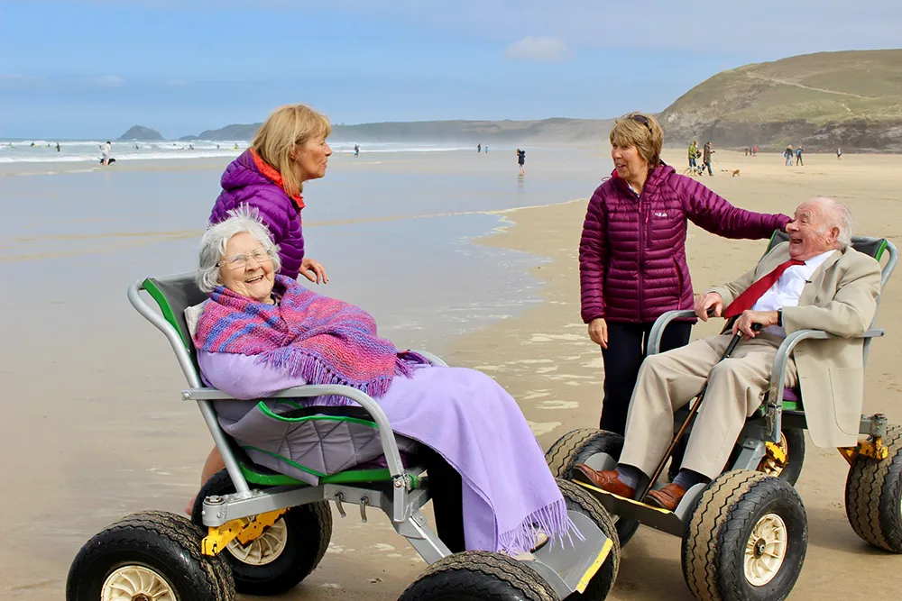 Two wheelchair users and their care workers smiling on the beach